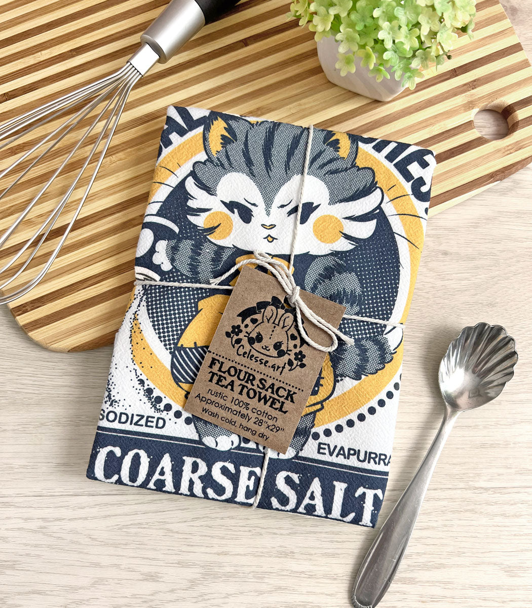 A folded tea towel tied with twine on a kitchen counter with a striped cutting board and some utensils, the printed design featuring a vintage-style logo for Salty Britches Coarse Salt with a kitten wearing patchwork britches and petulantly pouring out a container of salt.