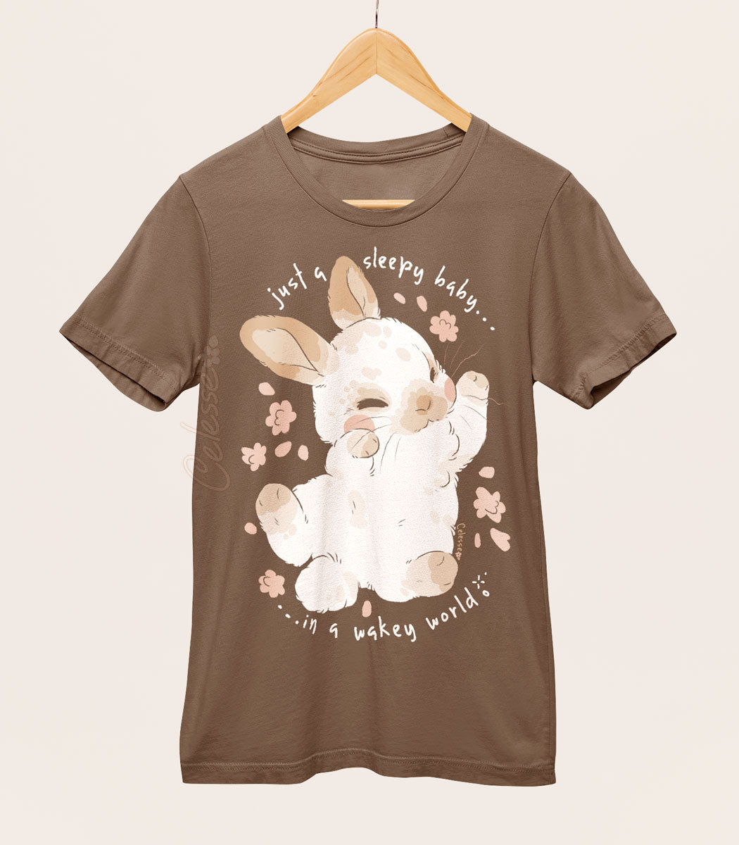 A brown shirt on a wood hanger with a print featuring a sleepy baby bunny sprawled out and the text "just a sleepy baby...in a wakey world."