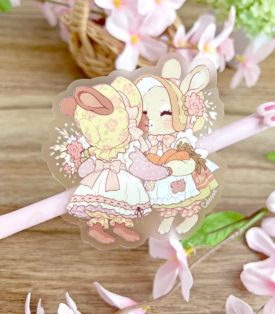 A Clear Vinyl Sticker of two bunnies wearing old-fashioned bonnets and dresses. One holds a bouquet, the other a bundle of carrots, and they lean in to share a soft kiss with each other.