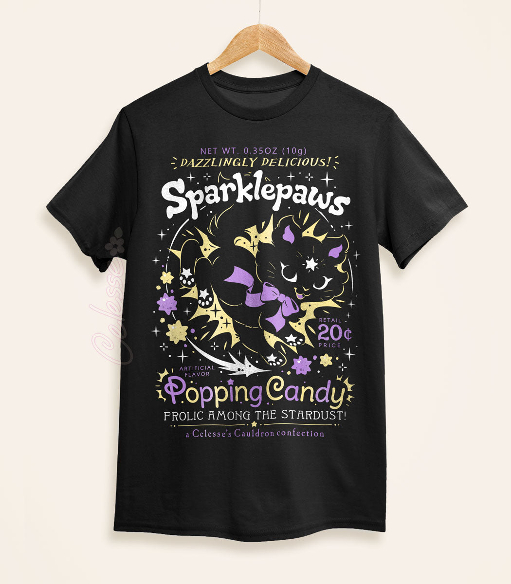 A black shirt on a wood hanger with a print of a retro faux candy logo for Sparklepaws Popping Candy, the art featuring a black cat with star paw pads leaping among bursting sparkles. The flavor text reads, "Dazzlingly delicious!" and "Frolic among the stardust!"