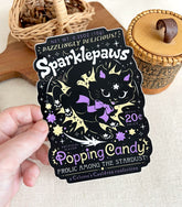 A hand holding a vinyl sticker with a print of a retro faux candy logo for Sparklepaws Popping Candy, the art featuring a black cat with star paw pads leaping among bursting sparkles. The flavor text reads, "Dazzlingly delicious!" and "Frolic among the stardust!"