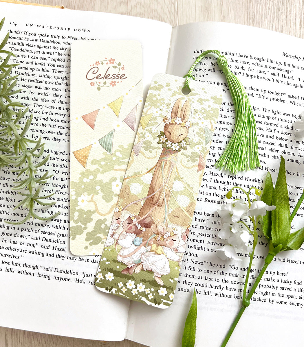 A photo of a bookmark placed atop an open book, the artwork featuring bunny girls in white dresses dancing around a maypole shaped like a rabbit.
