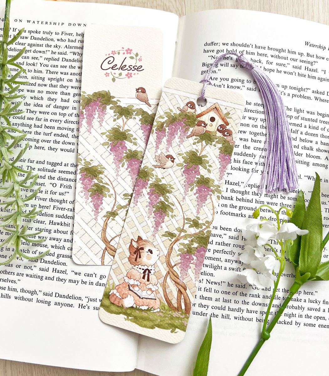 A photo of a bookmark placed atop an open book, the artwork featuring a kitten in a vintage dress with an open book in her lap, staring intently up at a birdhouse of chickadees atop a lattice wall of wisteria. 