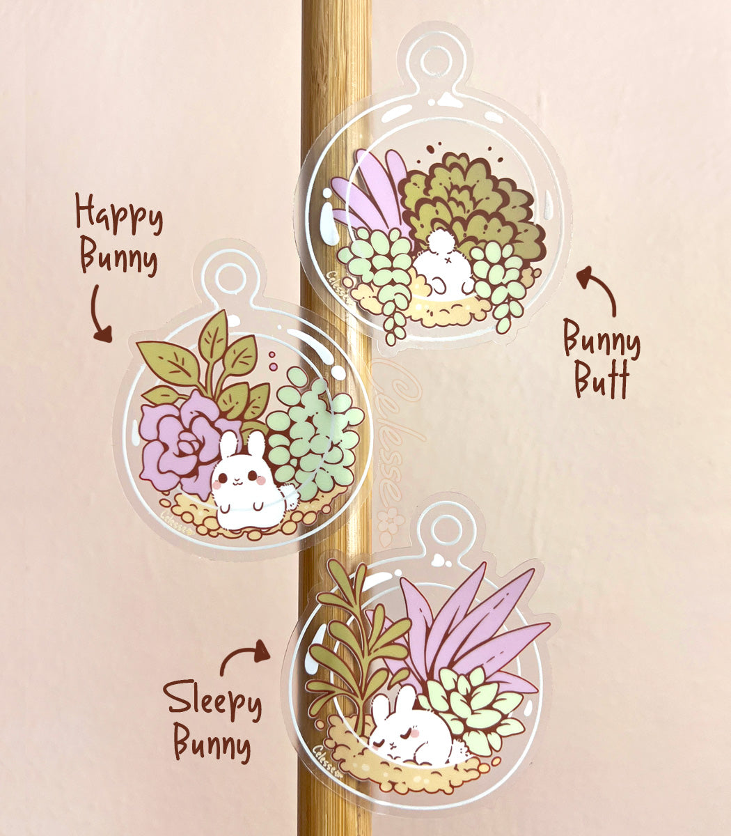 A photo of three clear vinyl stickers featuring art of tiny bunnies inside glass terrariums with succulents, stuck to a vertical wooden pole.