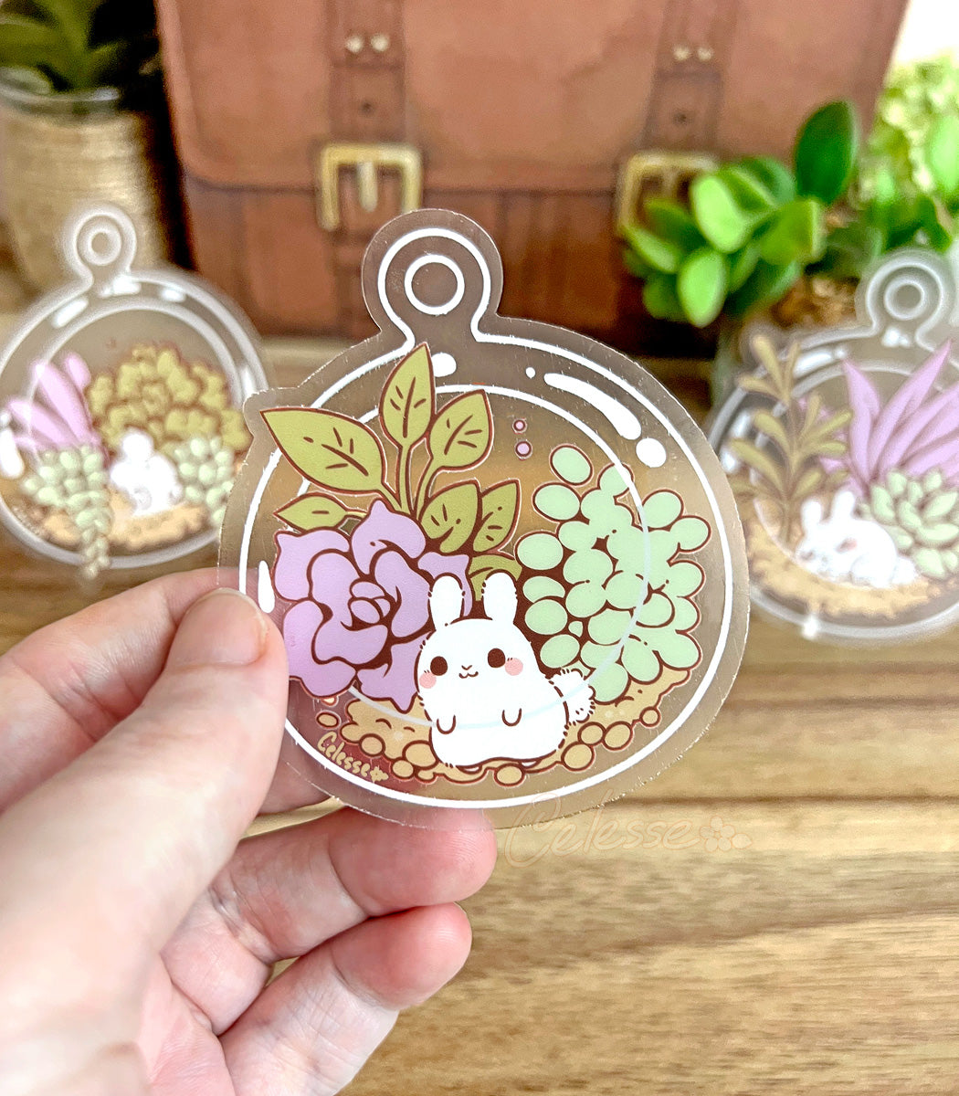 A photo of a hand holding a clear vinyl sticker featuring art of a tiny bunny inside a glass terrarium filled with succulents, a blurred wood table and plant decorations in the background.