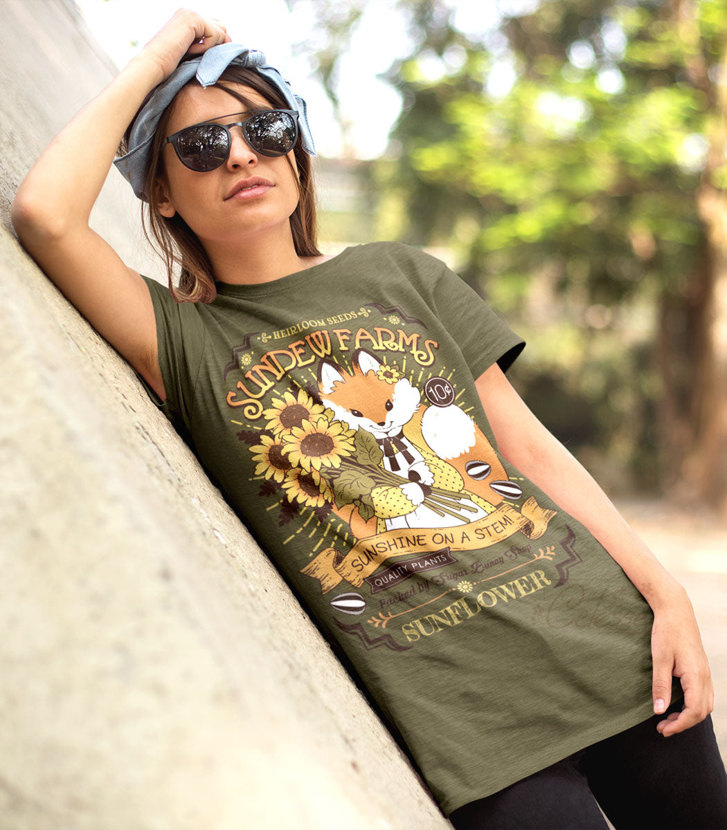 An angled outdoor shot of a person in sunglasses and leaning against a concrete wall, wearing an olive green shirt with a print of a vintage-style logo for Sundew Farms Sunflower Seeds that features a fox holding a bundle of sunflowers.