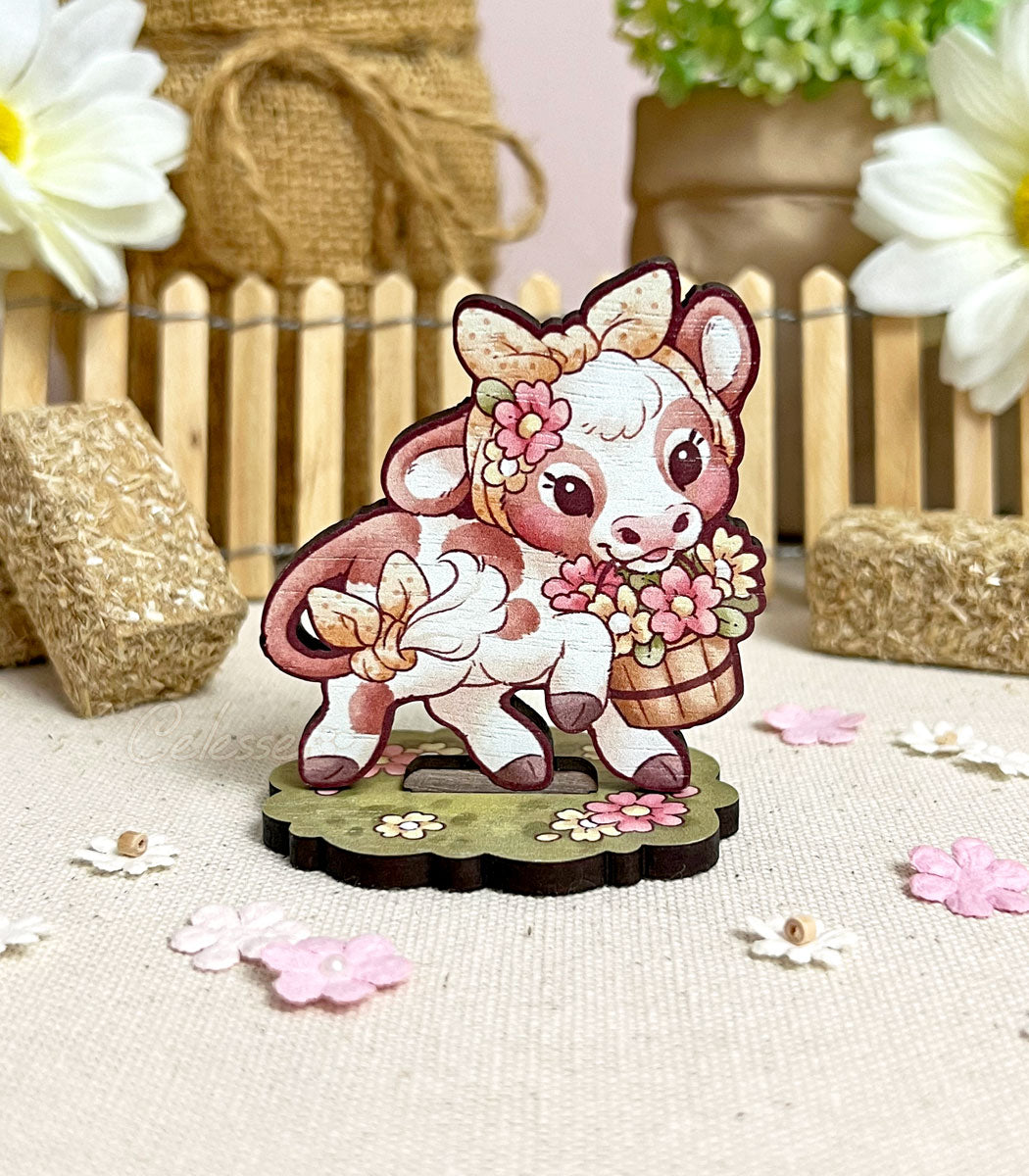A photo of a wooden standee featuring art of a baby cream-colored cow with light brown spots. She holds a bucket of flowers in her mouth and wears a yellow polka dot headband. Flowers, hay bales and a picket fence frame her in the background.