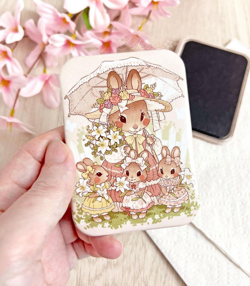 A hand holding a rectangular refrigerator magnet featuring artwork of a mother bunny in a vintage style dress holding a bundle of lilies and a lace parasol. Her three daughters stand in front of her each holding their own lily. The back side of a magnet is in the background.
