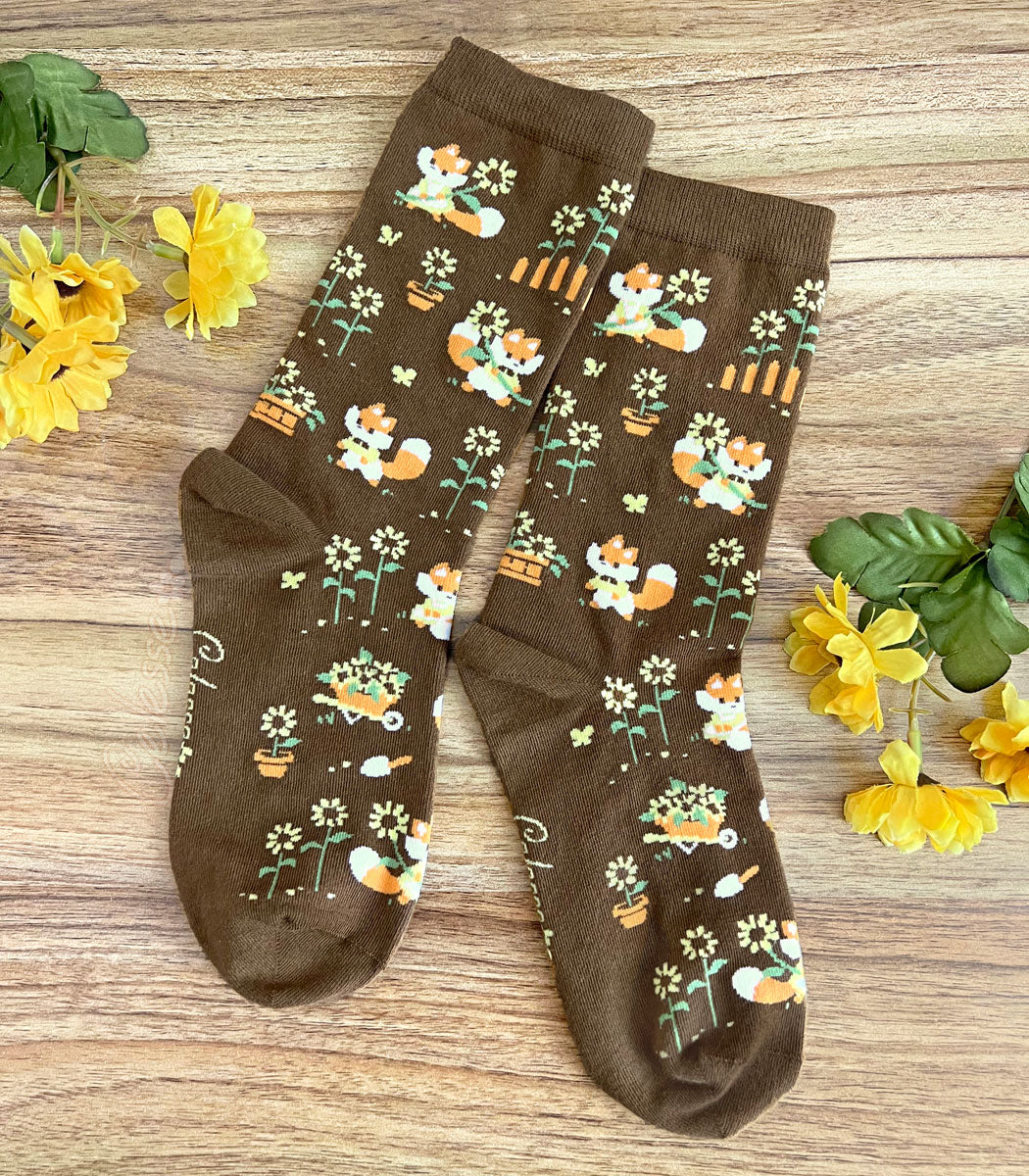 A pair of brown crew socks on a wood surface with flowers, the knit design featuring tiny pixel foxes in vintage clothes frolicking among sunflowers.