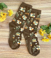 A pair of brown crew socks on a wood surface with flowers, the knit design featuring tiny pixel foxes in vintage clothes frolicking among sunflowers.