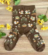 A pair of brown crew socks on a wood surface with flowers, the knit design featuring tiny pixel foxes in vintage clothes frolicking among sunflowers.