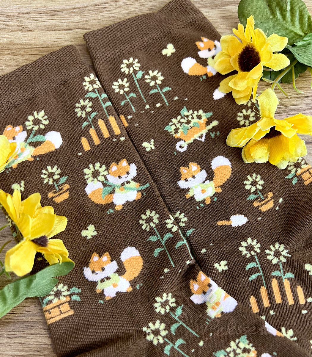 A close up of the knit details of a pair of brown crew socks on a wood surface with flowers, the design featuring tiny pixel foxes in vintage clothes frolicking among sunflowers.