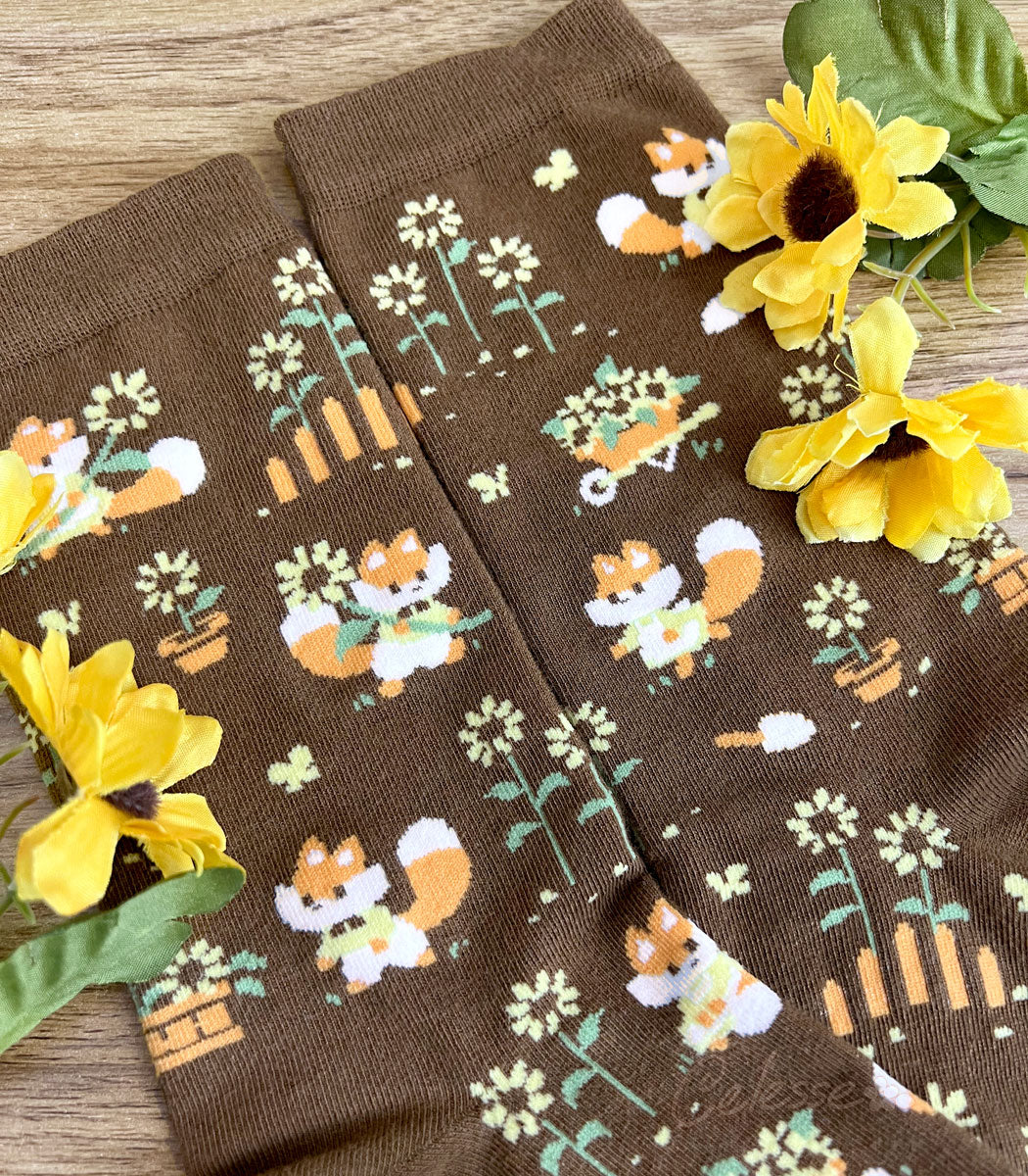 A close up of the knit details of a pair of brown crew socks on a wood surface with flowers, the design featuring tiny pixel foxes in vintage clothes frolicking among sunflowers.