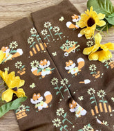 A close up of the knit details of a pair of brown crew socks on a wood surface with flowers, the design featuring tiny pixel foxes in vintage clothes frolicking among sunflowers.