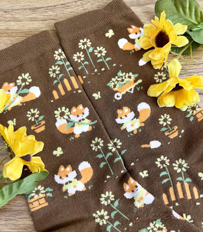 A close up of the knit details of a pair of brown crew socks on a wood surface with flowers, the design featuring tiny pixel foxes in vintage clothes frolicking among sunflowers.