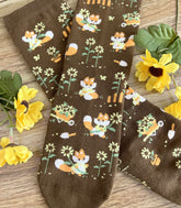 A pair of brown crew socks on a wood surface with flowers, the knit design featuring tiny pixel foxes in vintage clothes frolicking among sunflowers.