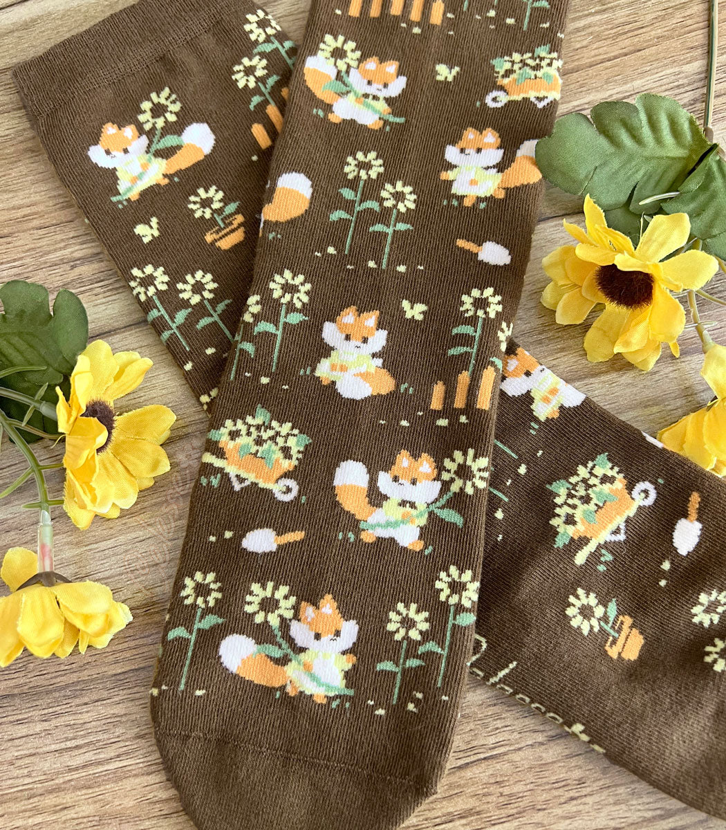 A pair of brown crew socks on a wood surface with flowers, the knit design featuring tiny pixel foxes in vintage clothes frolicking among sunflowers.