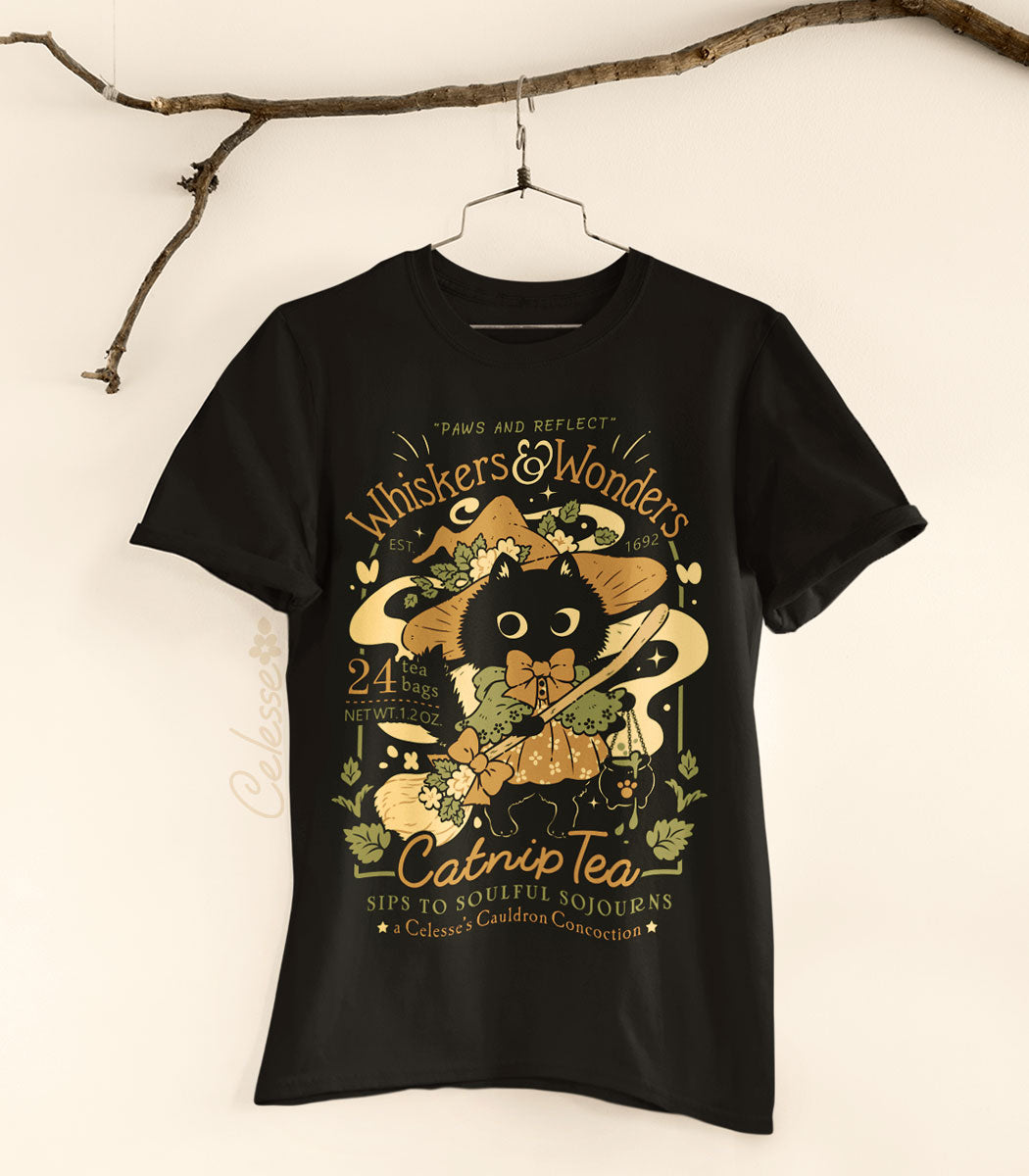 A photo of a shirt hung from a branch featuring a black cat witch holding a broom and a small bubbling cauldron, the text reads Whiskers &amp; Wonders Catnip Tea.