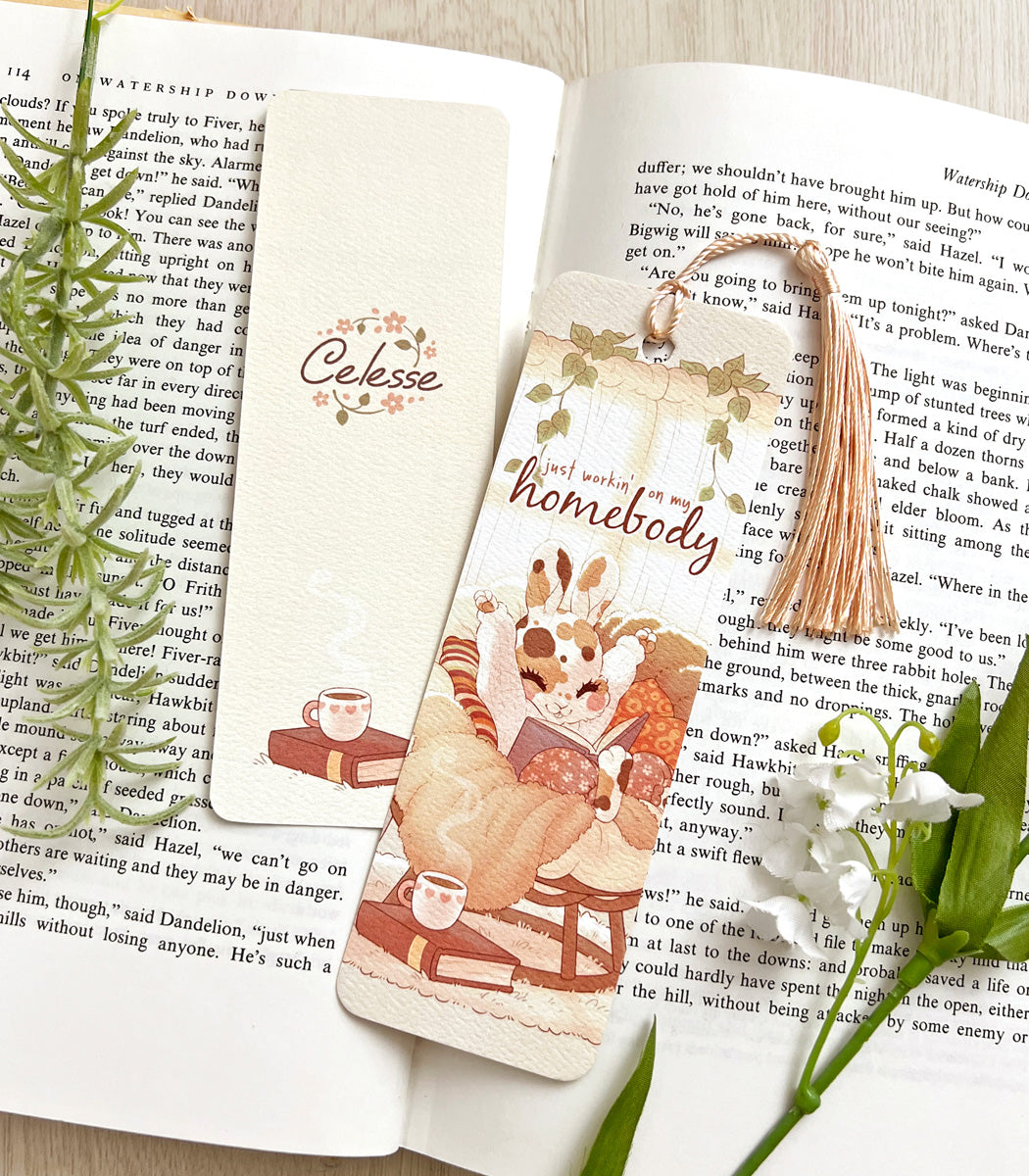 A photo of a bookmark placed atop an open book, the artwork featuring a bunny in a cozy sweater and snuggled up in a comfy papasan with a good book. The text reads "just workin&
