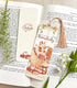 A photo of a bookmark placed atop an open book, the artwork featuring a bunny in a cozy sweater and snuggled up in a comfy papasan with a good book. The text reads "just workin&