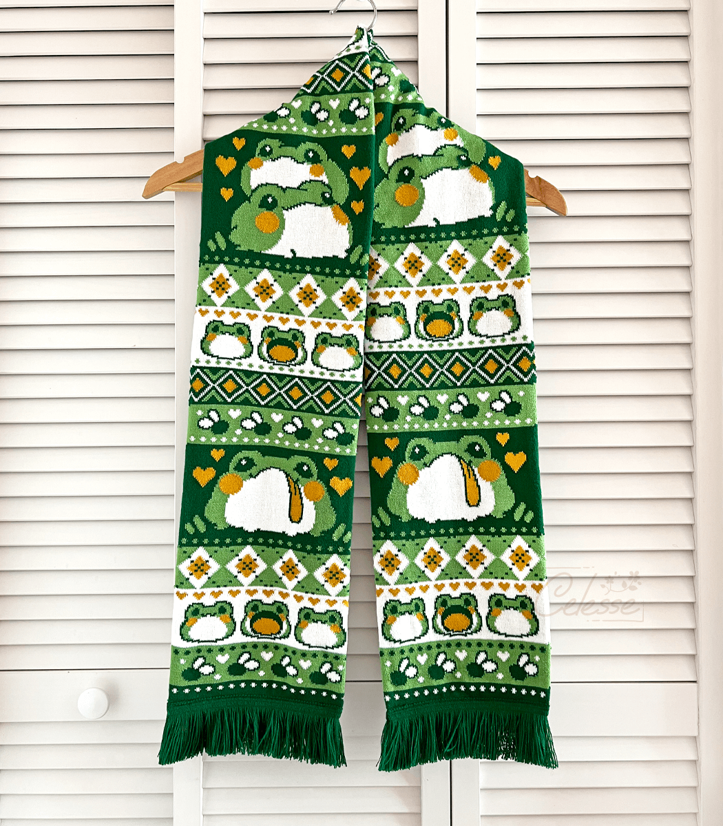 A photo of a knit scarf featuring fat blob frogs. The scarf is green, white and orange and has repeating knit patterns like diamonds, dots and flies.