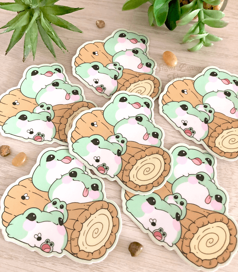 A photo of several vinyl stickers featuring cute, fat blob frogs, laid out on a wooden surface with plants in the background.