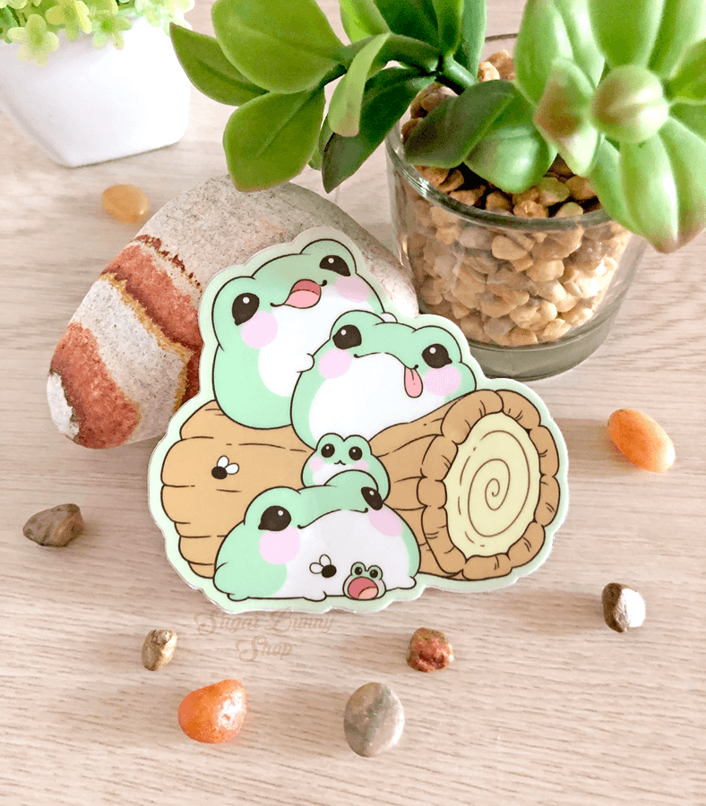 A vinyl sticker with a design of cute, fat green blob frogs, displayed on a wooden surface with a potted plant in the background.