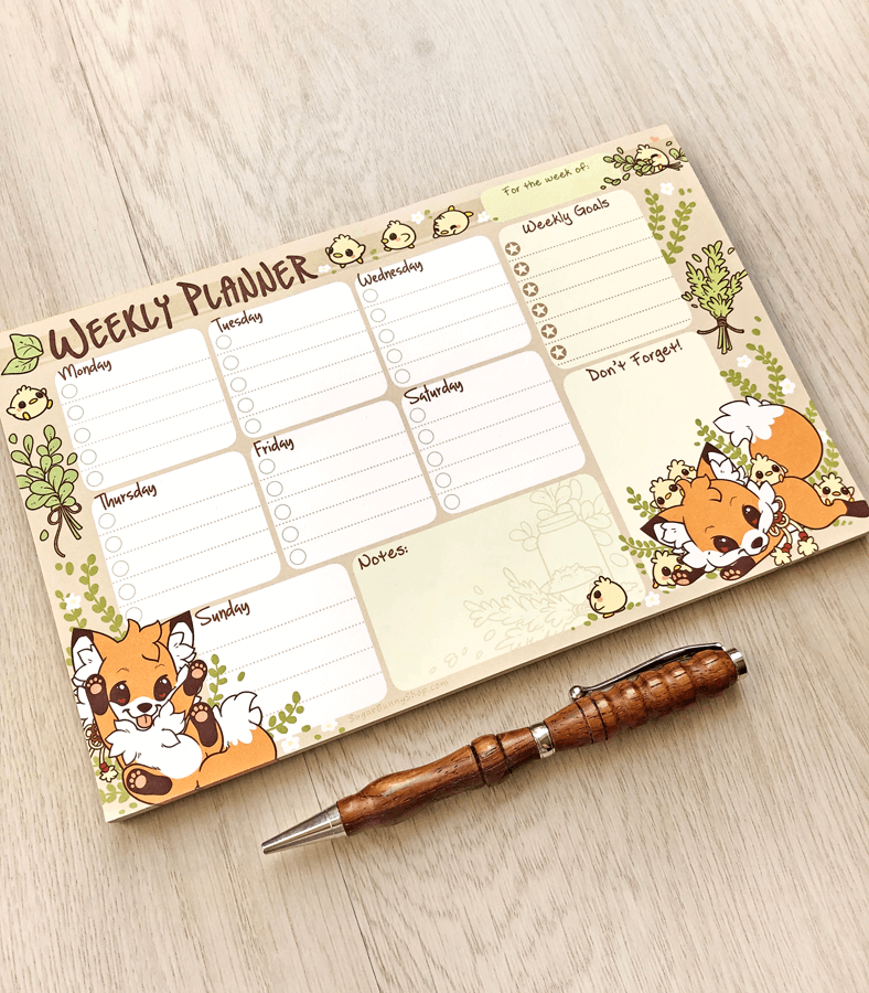 A photo of a weekly planner notepad on a wood table, the design featuring cute little foxes playing with yellow chicks, with task sections for each day of the week as well as extra notes and goals.