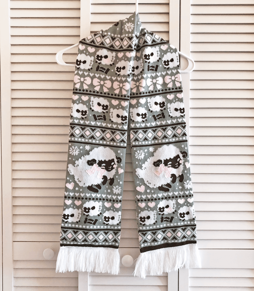 A photo of a knit scarf hung from slat wall doors, featuring fat fluffy white sheep with dark faces. The scarf is gray and white and has repeating knit patterns like diamonds, dots and hearts.