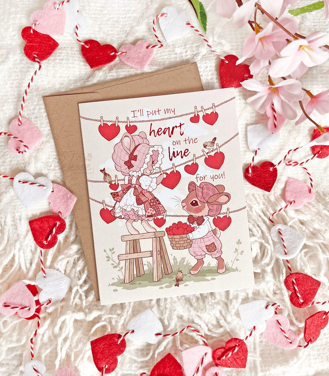 A photo of a vintage-style greeting card featuring soft artwork of two bunnies hanging paper hearts on a clothesline. The text reads "I&