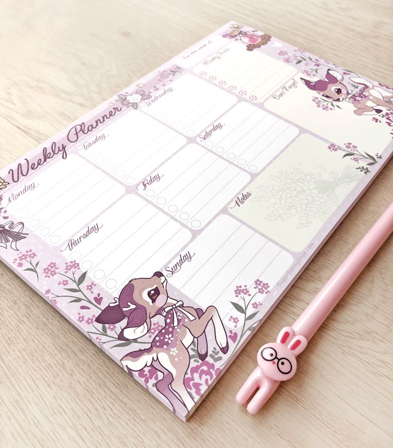 A photo of a weekly planner notepad on a wood table, the design featuring a lavender theme with cute baby deer frolicking around task sections for each day of the week, as well as extra sections for notes and goals.