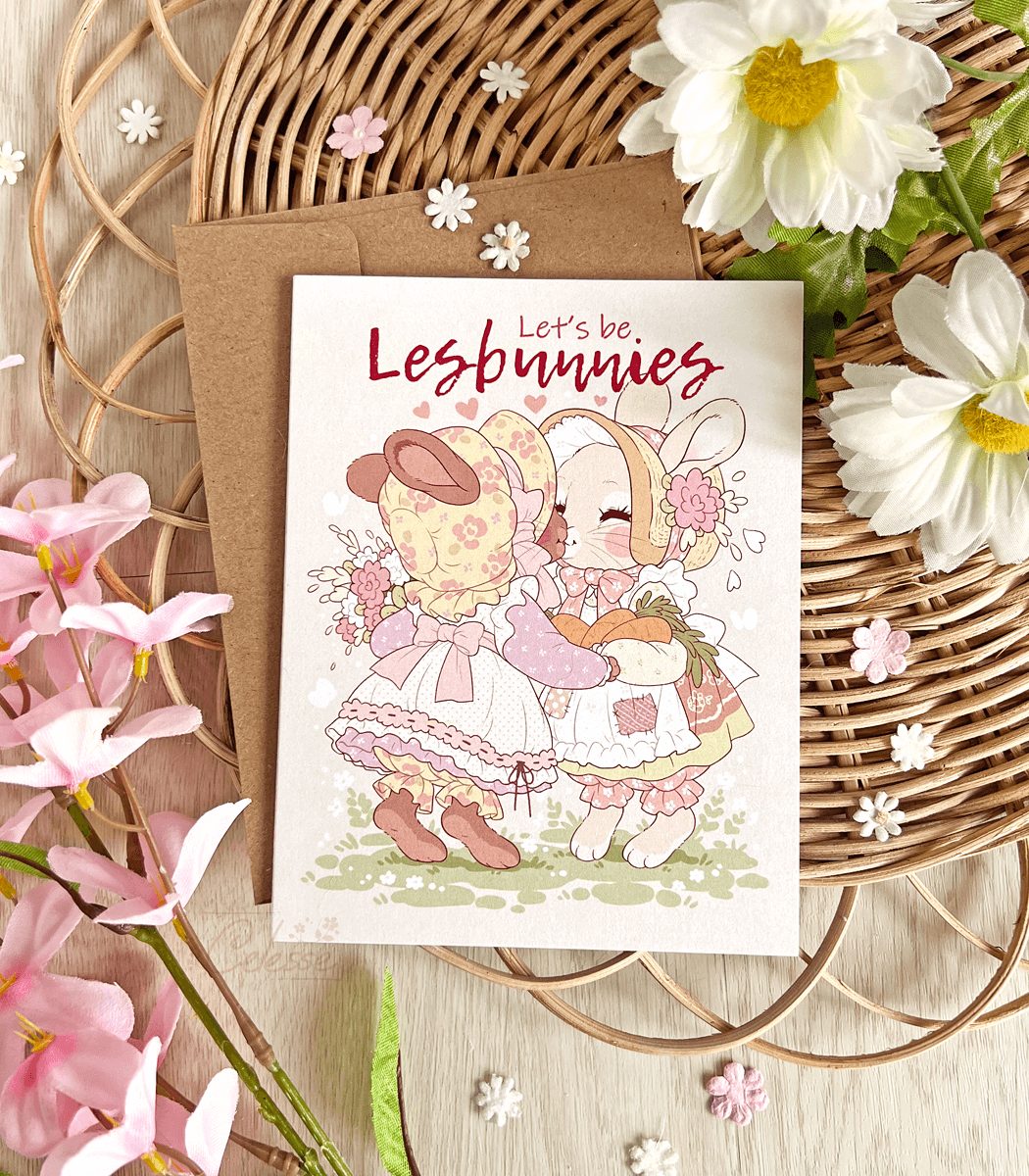 A photo of a vintage-style greeting card featuring soft artwork of two bunnies wearing bonnets and floral dresses sharing a kiss. The text reads "Let&