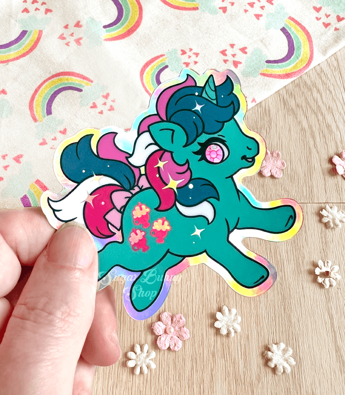 A photo of a hand holding a holographic vinyl sticker featuring Fizzy the unicorn from the first generation of My Little Pony. She has a green body, multicolored hair of dark green, white and magenta, and has milkshakes for a cutie mark. A rainbow cloth and tiny flowers decorate the background.