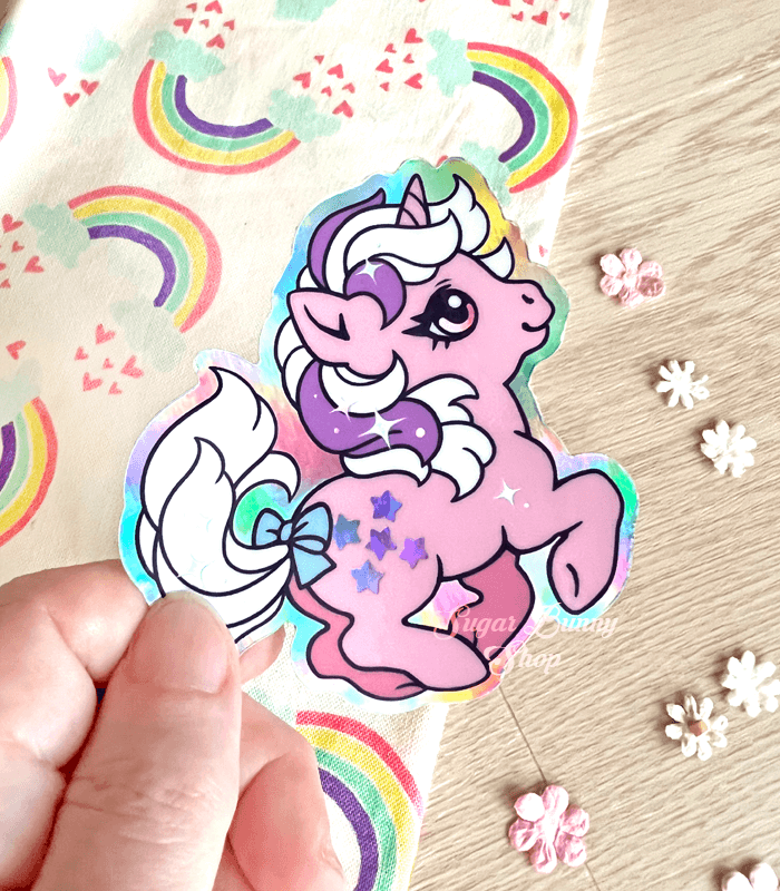 A photo of a hand holding a holographic vinyl sticker featuring Twilight the unicorn from the first generation of My Little Pony. She has a pink body, white hair with a purple stripe and stars for a cutie mark. A rainbow cloth and tiny flowers decorate the background.