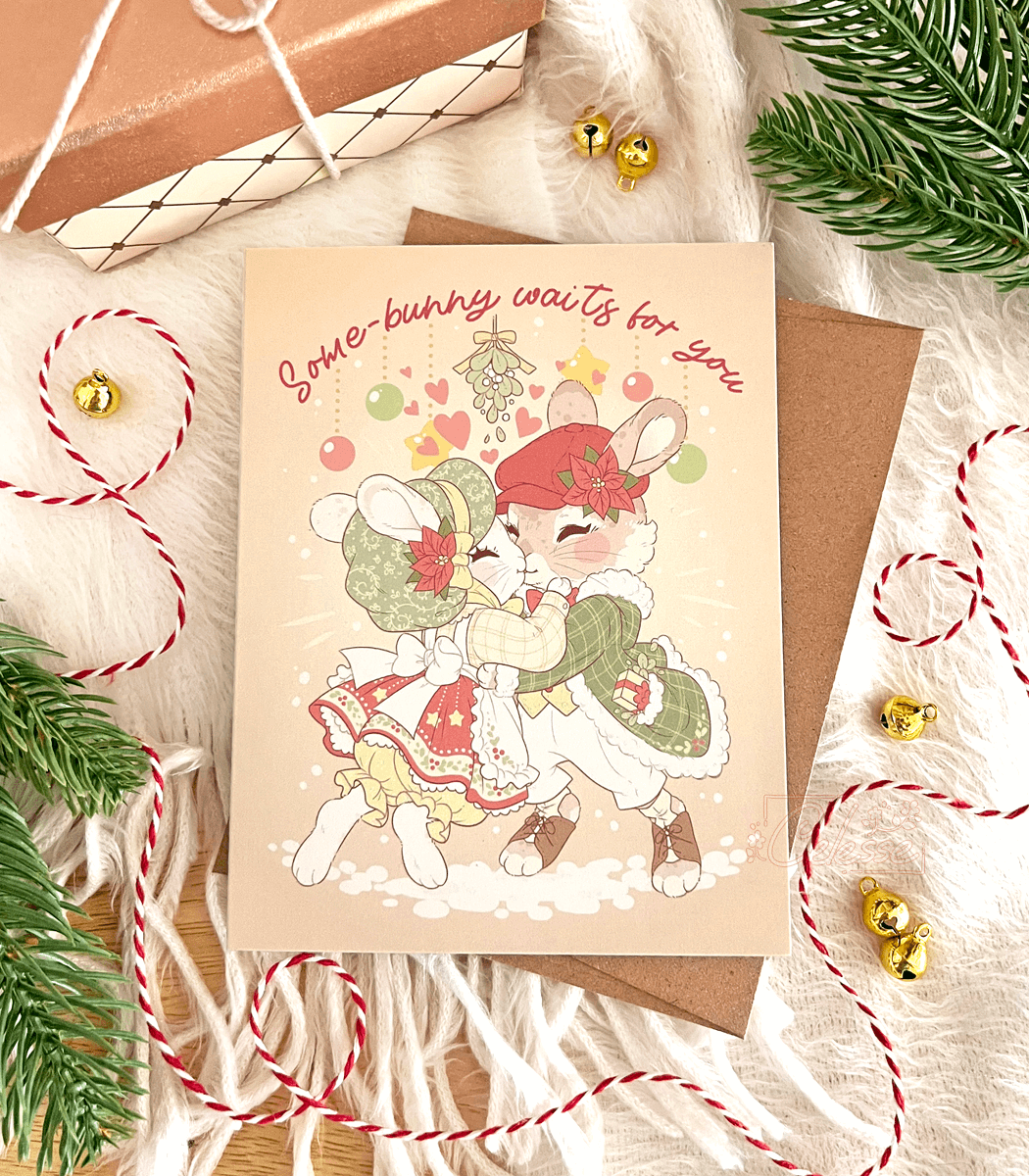 A photo of a Christmas greeting card featuring vintage style artwork of two bunnies dressed in old-fashioned winter clothing and happily meeting for a kiss under the mistletoe. The text reads "Some-bunny waits for you" An envelope, string and other decorations frame it.