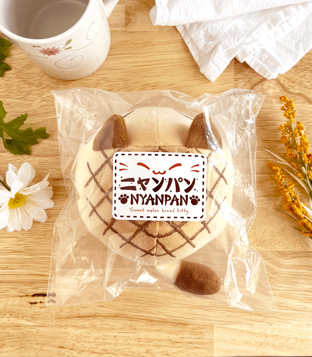 A round plush bread cat in the shape of melon bread inside plastic packaging on a kitchen counter, with a "Nyanpan" label on top.