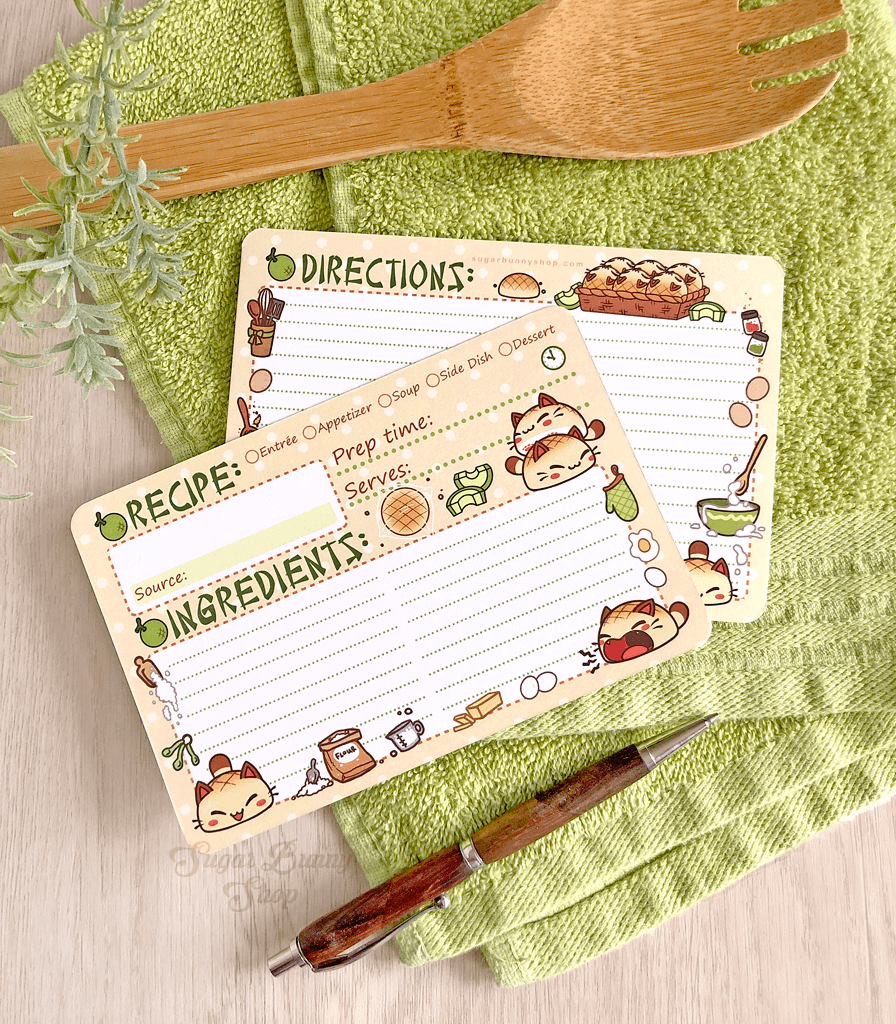 A set of recipe cards with an illustration of little bread cats and kitchen elements, placed on a green kitchen towel alongside a wooden spoon and a pen.