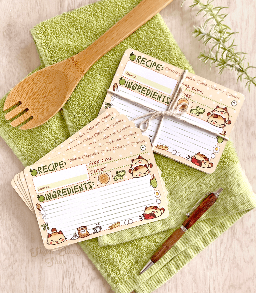 A set of recipe cards with an illustration of little bread cats and kitchen elements, placed on a green kitchen towel alongside a wooden spoon and a pen.