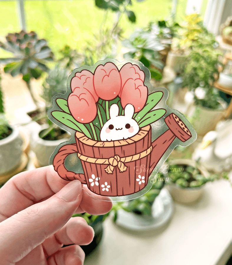 A hand holding a clear vinyl sticker in front of a bunch of plants, the art featuring a tiny bunny nestled in a wood watering can with tulips growing out of it.