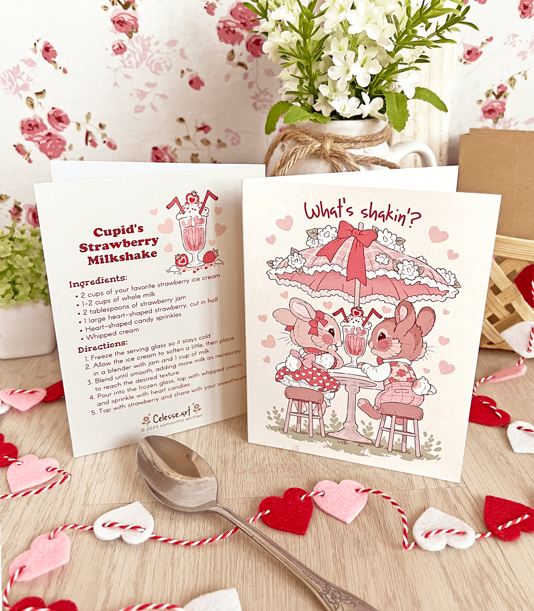 A photo of a vintage-style greeting card featuring soft artwork of two bunnies sitting at a table with a parasol and sharing a strawberry milkshake. The text on the front reads "What's shakin'?" and the back features a recipe for a strawberry milkshake.